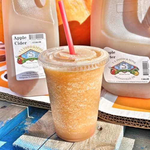 A clear cup of apple cider slush sits on a table, surrounded by pumpkins and other cider containers.