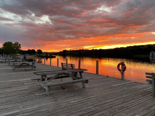 A serene lakeside view at sunset, with vibrant orange skies reflecting on the water and wooden picnic tables on the dock.
