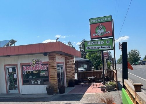 A sunny street view of a restaurant with outdoor seating, featuring signs for breakfast, bistro, and cocktails.