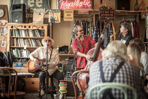 Three musicians perform in a cozy café, with a guitarist, a bassist, and a singer, while an audience enjoys the show.