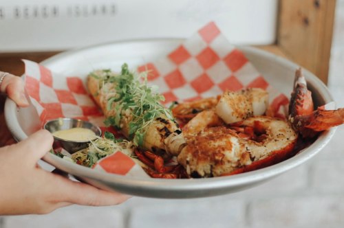 A hand holds a metal platter with grilled seafood, including shrimp, a stuffed roll, and garnished with greens.