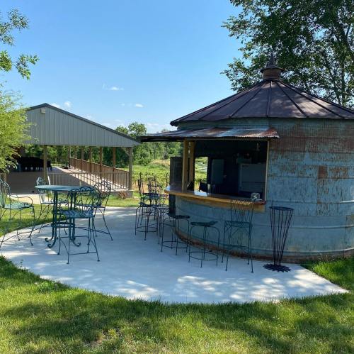 A rustic outdoor bar with metal walls, surrounded by green grass and trees, featuring tables and chairs.