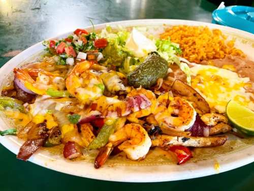 A colorful plate of sizzling shrimp and vegetables topped with cheese, served with rice, beans, and fresh toppings.
