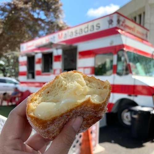 A hand holding a half-eaten malasada, with a food truck in the background featuring red and white stripes.