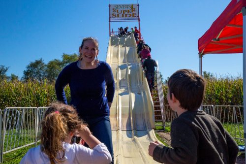 A woman smiles at children near a large slide at a fair, with a cornfield in the background and kids waiting to slide.
