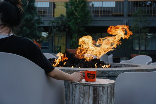A person sits by a fire pit, holding an orange mug, with flames dancing in the background.