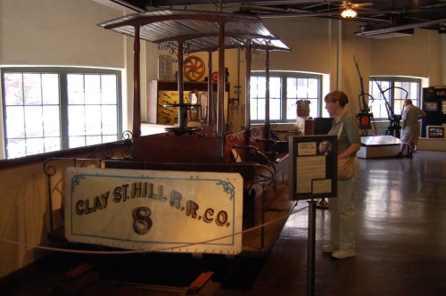 A vintage streetcar on display in a museum, with a visitor examining it and various artifacts in the background.