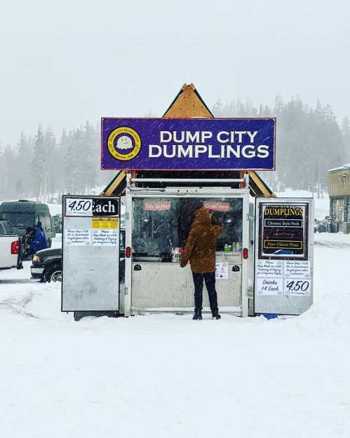 A person stands at a food stall named "Dump City Dumplings" in a snowy landscape, with a sign displaying menu prices.