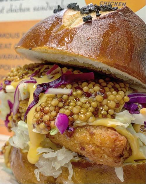 Close-up of a pretzel bun sandwich filled with fried chicken, mustard seeds, cabbage, and a drizzle of yellow sauce.