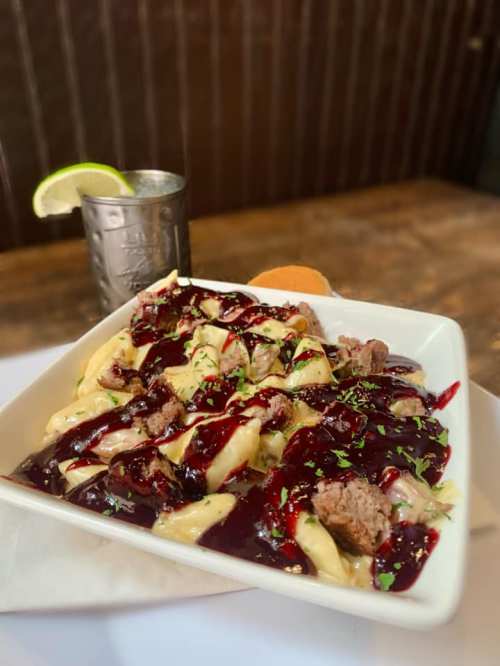 A plate of pasta topped with meat, drizzled in a dark sauce, garnished with herbs, alongside a drink with a lime wedge.