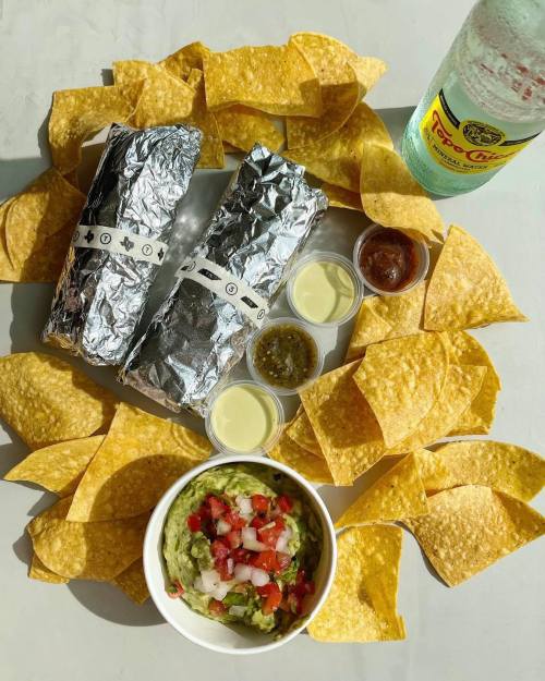 A spread of two wrapped burritos, guacamole with toppings, sauces, and tortilla chips, surrounded by a bottle of soda.