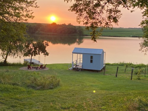 A serene lakeside scene at sunset, featuring a small cabin, chairs, and lush green grass.