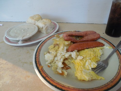 A plate of scrambled eggs, sausage, and hash browns, with biscuits and gravy on the side, and a drink.
