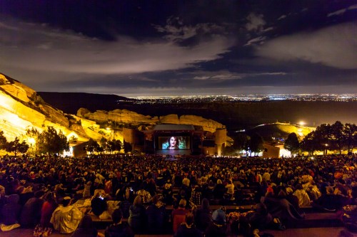 A large crowd watches a movie on a big screen at night, surrounded by illuminated rock formations and city lights.