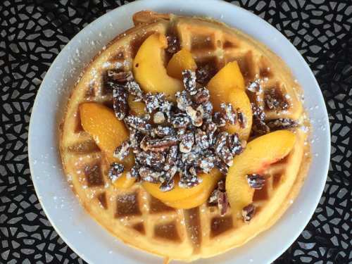 A golden waffle topped with sliced peaches, pecans, and a dusting of powdered sugar on a decorative plate.