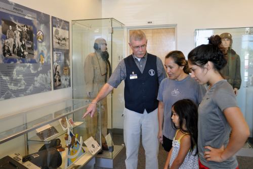 A museum guide explains exhibits to a woman and two girls, with displays of historical items in the background.