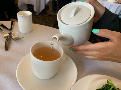 A hand pours tea from a white teapot into a white cup on a table set with utensils.