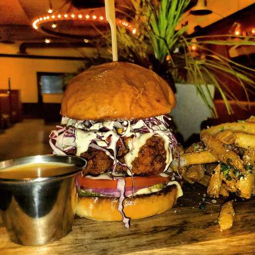 A close-up of a burger with fried chicken, lettuce, and sauce, served with a side of fries and dipping sauce.