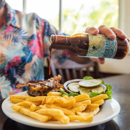 A person in a colorful shirt pours sauce over a burger and fries on a plate at a restaurant.