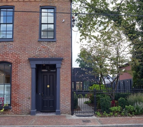 A brick building with a black door and windows, surrounded by greenery and a fence, on a quiet street.