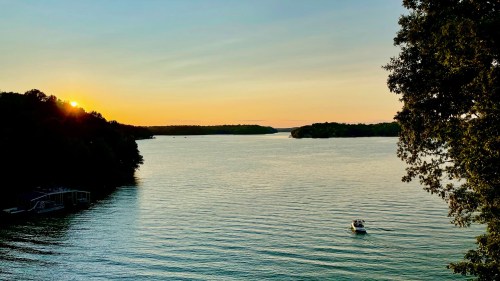 A serene sunset over a calm lake, with a small boat gently gliding through the water and trees lining the shore.