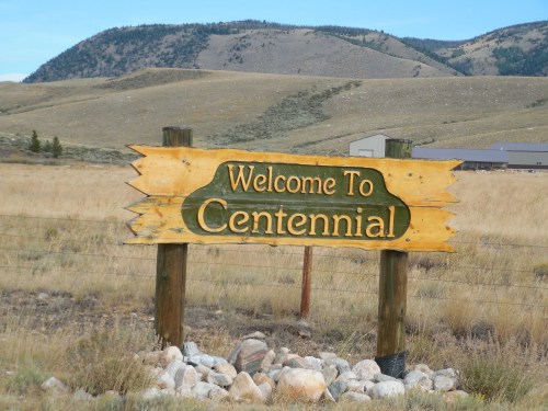 Wooden sign reading "Welcome to Centennial" in a grassy landscape with mountains in the background.