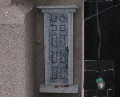 A small, decorative door sculpture mounted on a wall, with coins and a rose placed at its base.