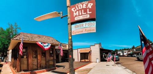 A rustic cider mill with a sign, flags, and a clear blue sky, showcasing a charming small-town atmosphere.