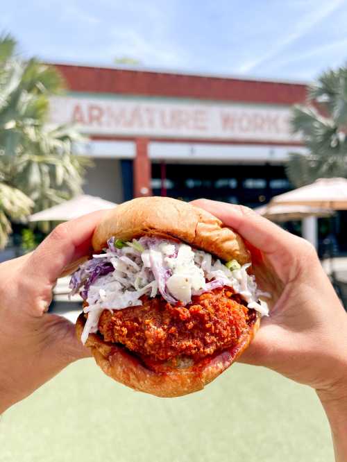 A person holds a crispy fried chicken sandwich topped with coleslaw, set against a sunny outdoor backdrop.