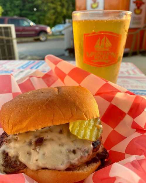 A close-up of a cheeseburger with pickles on a red-checkered plate, alongside a glass of beer.