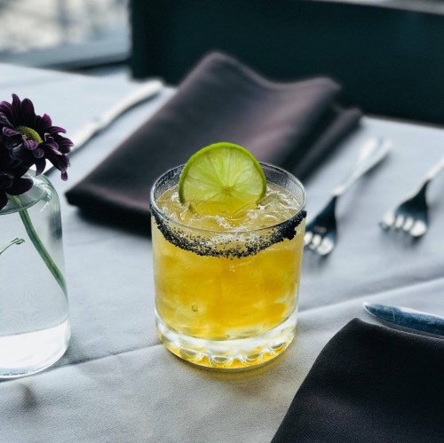A refreshing cocktail with a lime slice, served in a glass with a black salt rim, on a table with a flower vase.
