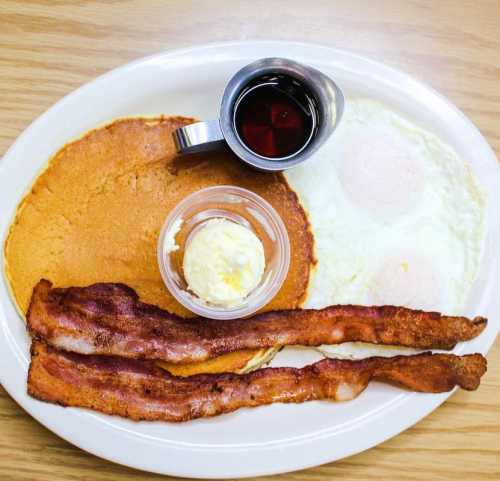 A plate with pancakes, two fried eggs, crispy bacon, a small cup of butter, and a syrup container.