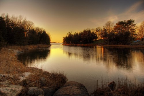 A serene river at sunset, reflecting warm colors, surrounded by trees and rocky banks.