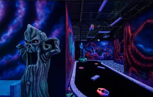 A vibrant indoor mini-golf course featuring glowing murals and a spooky stone statue.