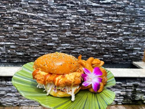 A sesame seed bun sandwich with fried chicken, slaw, and a flower, served with curly fries on a green leaf.