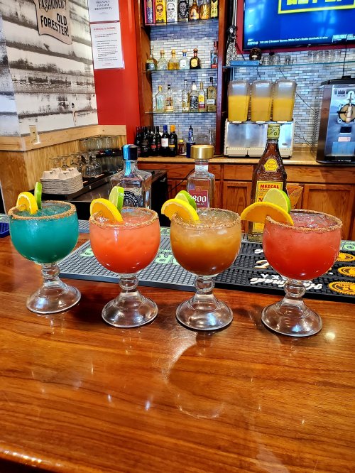 Four colorful cocktails with garnishes of lime and orange sit on a wooden bar, showcasing vibrant hues and salt rims.