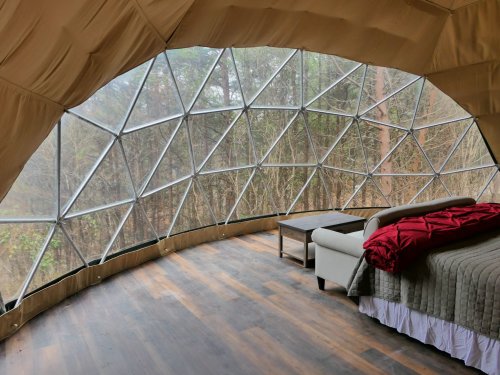 Interior of a geodesic dome with a bed, sofa, and large windows overlooking a forested area.
