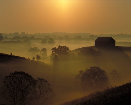 A serene landscape at sunrise, featuring rolling hills, a silhouette of a barn, and misty trees.