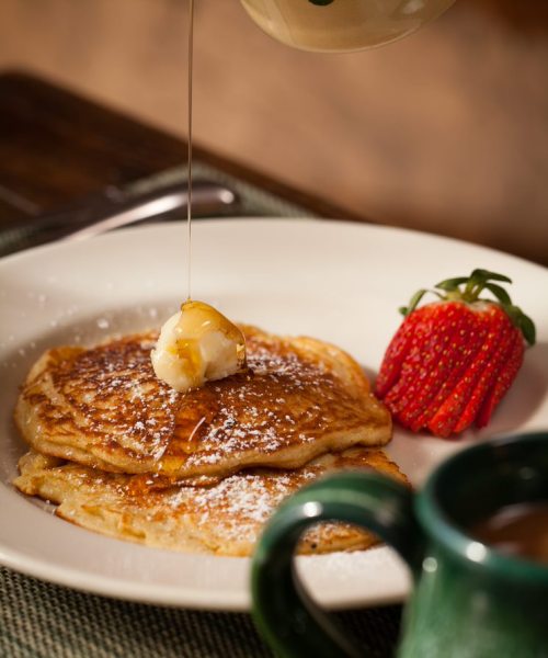 A stack of pancakes topped with melting butter and syrup, accompanied by a fresh strawberry on a white plate.
