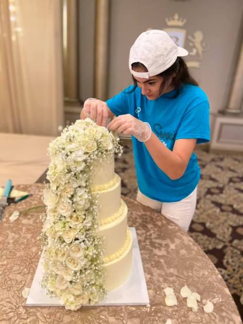A person decorates a three-tiered cake with white flowers and greenery, wearing gloves and a blue shirt.