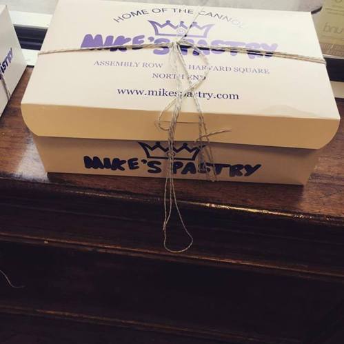 Two pastry boxes tied with string, labeled "Mike's Pastry" and "Home of the Cannoli," on a wooden surface.