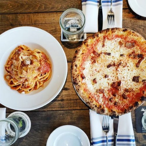 A wooden table with a plate of spaghetti and a pizza, accompanied by glasses and utensils.
