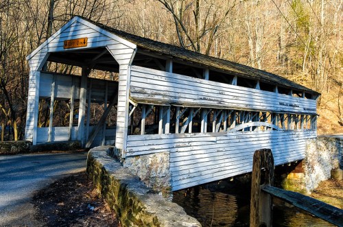A weathered covered bridge spans a small stream, surrounded by bare trees and a rocky bank.