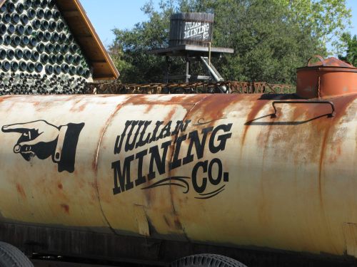 Rusty tank with "Julian Mining Co." logo and a pointing hand, set against a backdrop of trees and a wooden structure.