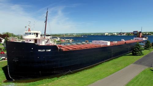 A large black ship named "Valley Camp" docked by a waterfront, surrounded by green grass and blue skies.