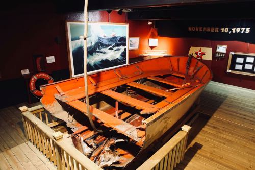 A damaged orange lifeboat displayed in a museum, surrounded by wooden flooring and informational exhibits.