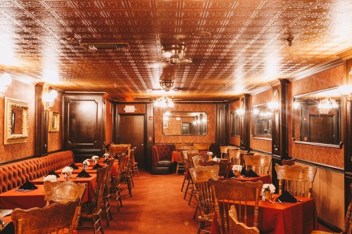 Elegant restaurant interior with ornate ceiling, wooden accents, and tables set with black napkins and floral centerpieces.