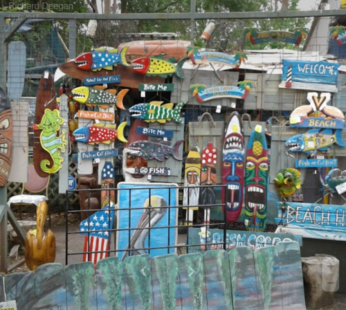 Colorful wooden signs and sculptures, including fish, lizards, and tiki faces, displayed in a vibrant outdoor setting.