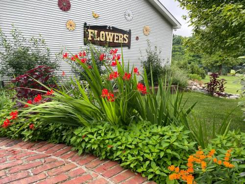 A vibrant flower garden with red and orange blooms, lush green plants, and a sign reading "Flowers" on a house.