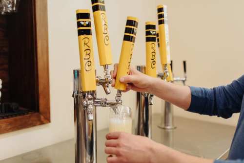A person pours a drink from a yellow-topped beer tap into a glass at a bar.